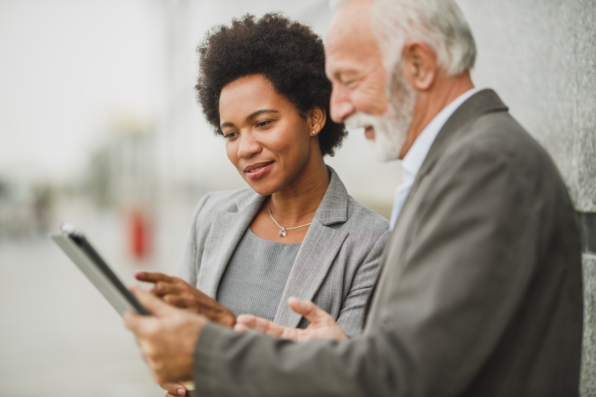 Shot of successful black businesswoman and her senior male colleague using digital tablet and having a discussion while standing against a wall of corporate building.